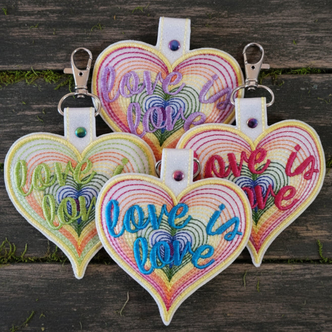 A colorful keychain with a heart shape and the phrase 'Love is Love' written in a stylized font, designed to resemble a well-known television show character's signature.