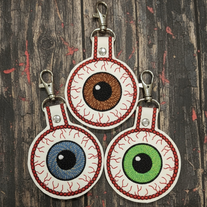 A collection of three handmade keychains shaped like eyes, with different colors for the irises, attached to a lobster clasp hook.