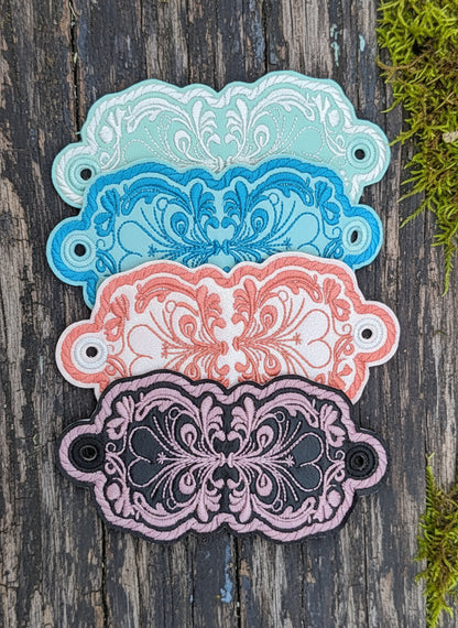 Three hair holders with floral filigree patterns in blue, pink, and white colors, made with vinyl and cotton stitching, displayed against a dark background.