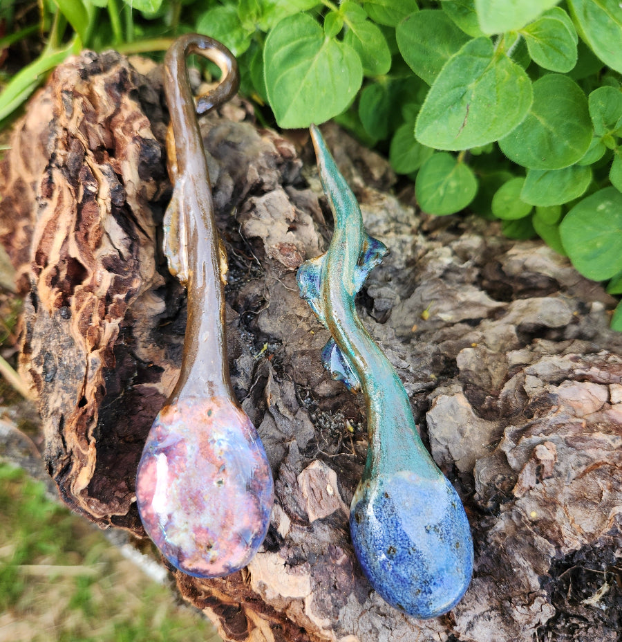 Two glass spoons, one pink and one blue, on a wooden surface with green leaves in the background.