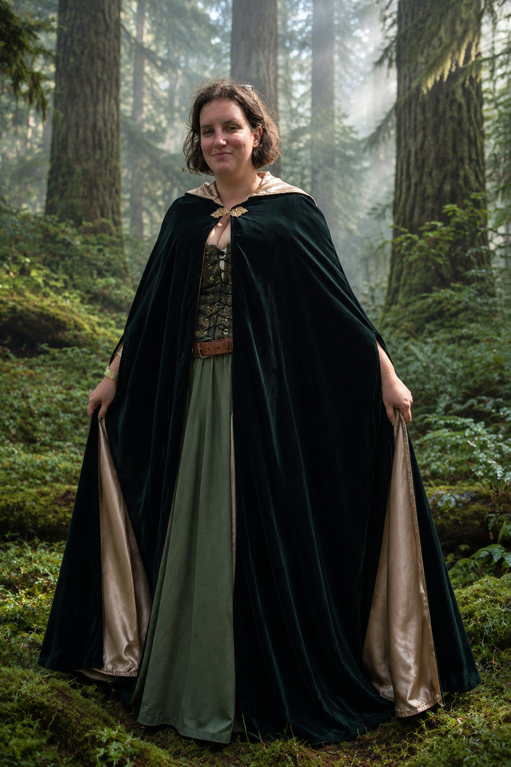 A woman wearing a medieval-style costume with a dark green cloak over a green dress, standing outdoors with  trees in the background.