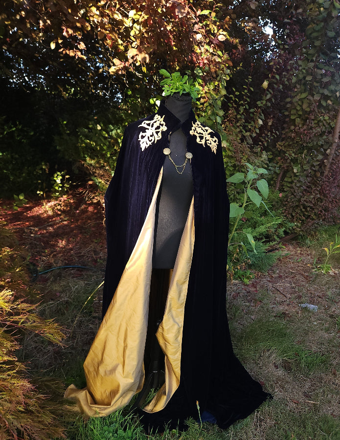 A long black velvet cape with ornate gold embroidery on the shoulders and a gold silk lining, displayed on a mannequin outdoors in a garden setting.