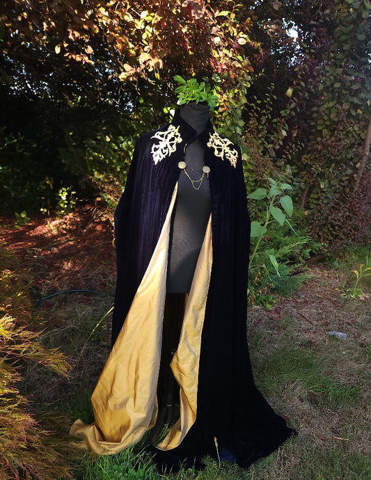 A long black velvet cape with ornate gold embroidery on the shoulders and a gold silk lining, displayed on a mannequin outdoors in a garden setting.