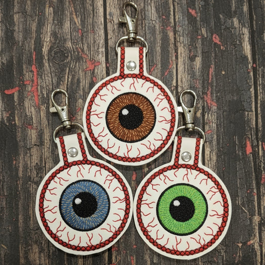 A collection of three handmade keychains shaped like eyes, with different colors for the irises, attached to a lobster clasp hook.