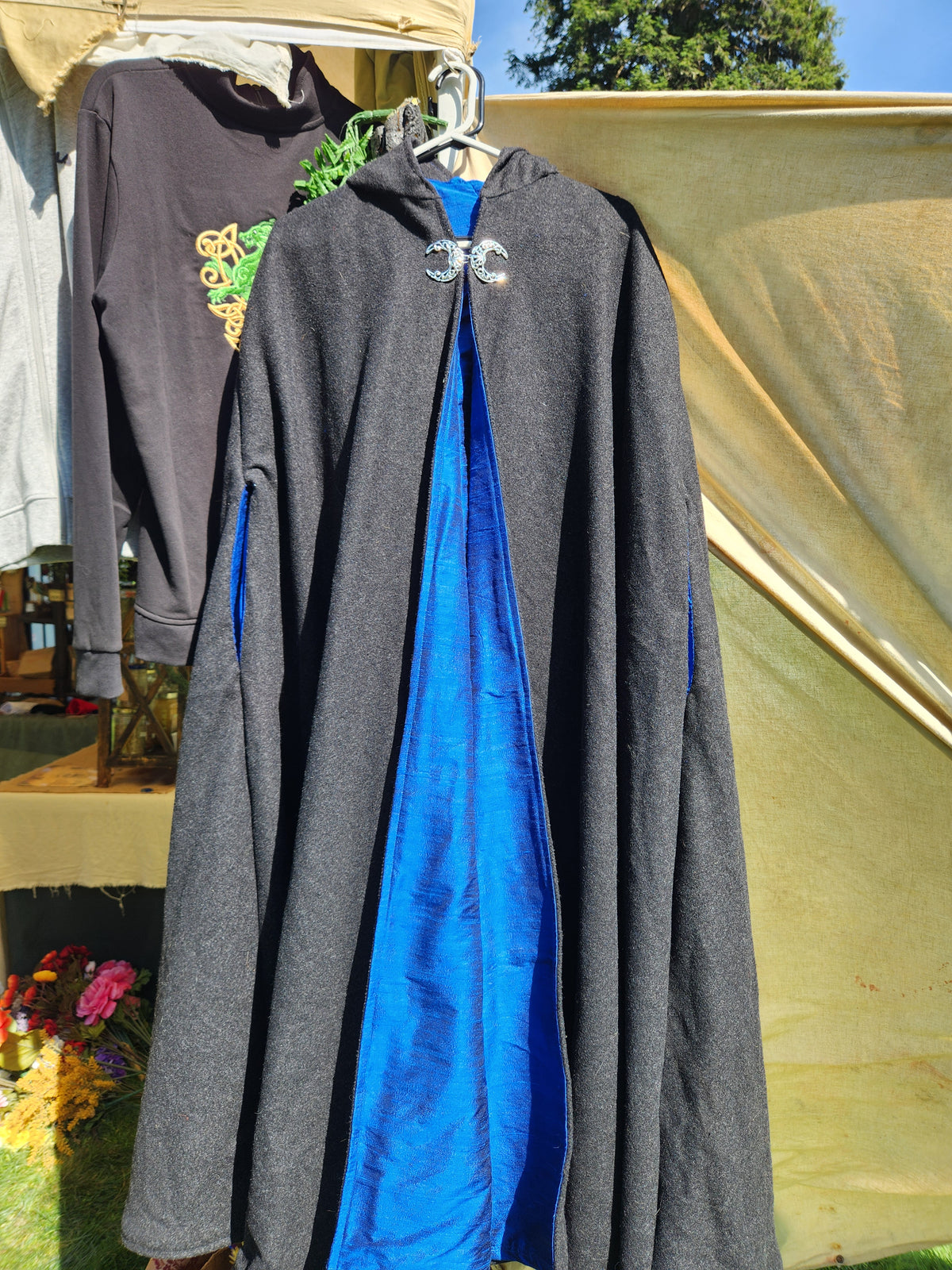A long dark gray hooded cloak or robe with a bright blue silk inner lining and a silver double moonemblem at the neckline, hanging outdoors against a beige tent backdrop.